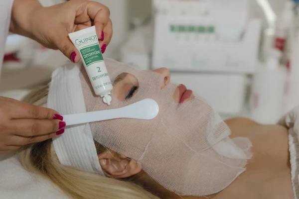 Therapist applying a detoxifying facial mask over a protective gauze layer during a professional spa facial treatment.