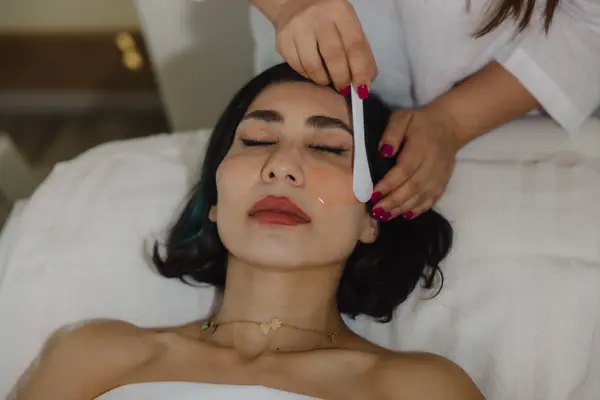 Woman relaxing on a treatment bed while a therapist gently applies skincare product to her face using a spatula.