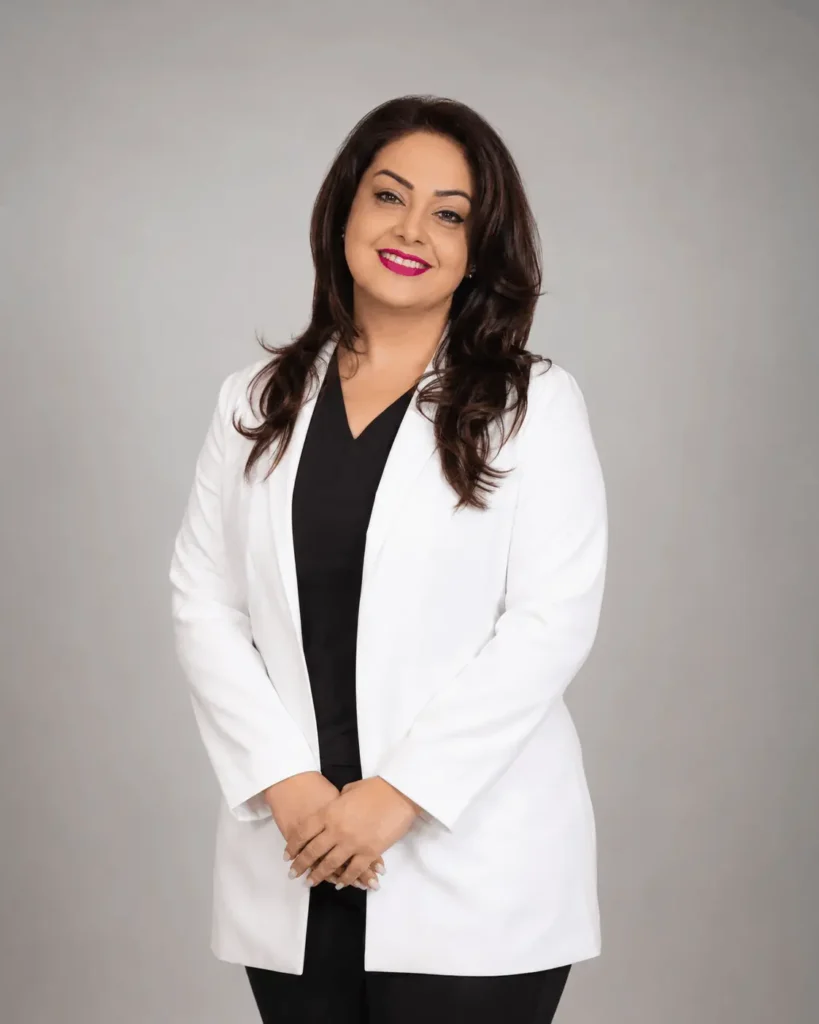 Professional studio portrait of a smiling woman with dark layered hair, wearing a white lab coat over a black V-neck top, standing with her hands gently clasped in front of a soft light-gray background.